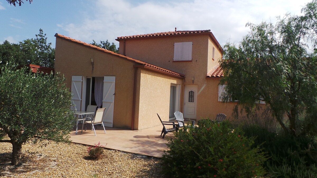 A single-story villa is positioned amid a landscaped garden, featuring beige exterior walls and a terracotta roof. Outdoor seating is visible, showing two chairs and a table on the wooden terrace. Lush greenery surrounds the property, with olive trees and shrubs enhancing the natural setting.