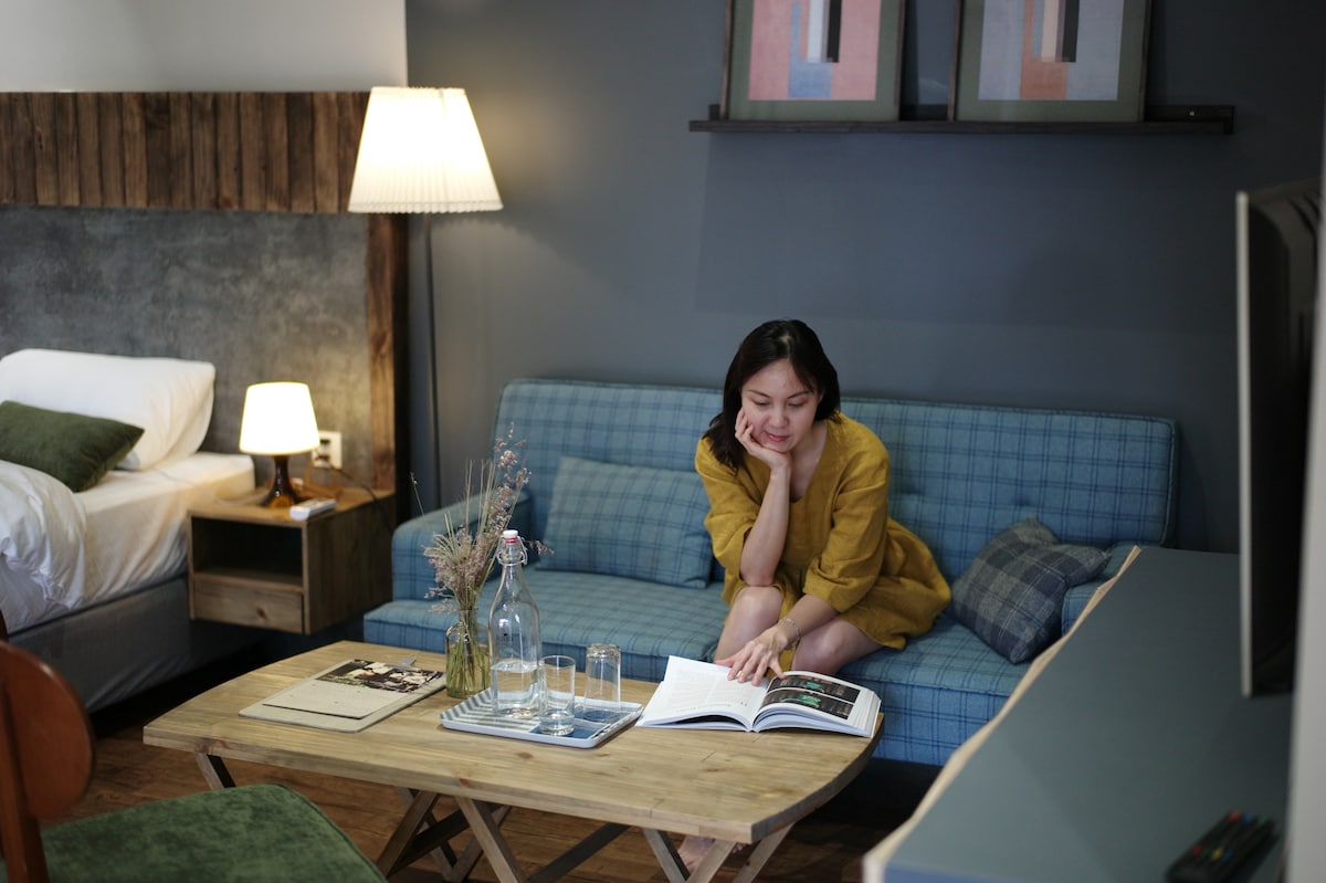 A cozy living area is presented with a plaid sofa and a wooden coffee table. A soft glow from a lamp beside the sofa adds warmth. A magazine and a glass bottle sit on the table. A bed is partially visible in the background.