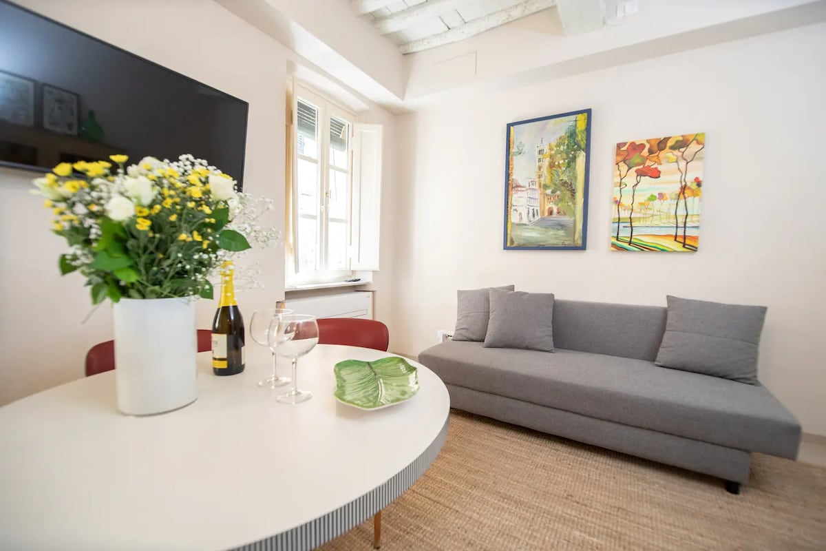 A light-filled living area features a gray sofa and a circular dining table. A floral arrangement in a white vase sits prominently on the table, accompanied by glasses and a bottle. Two colorful paintings adorn the walls, adding visual interest to the space.