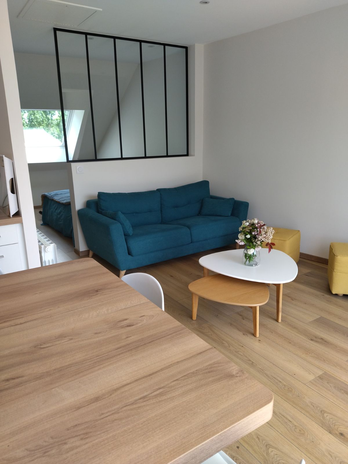 A bright and welcoming living area features a blue sofa, a round coffee table, and a vase with flowers. A small yellow armchair is positioned nearby. The interior design includes wooden flooring and a large window providing ample natural light.