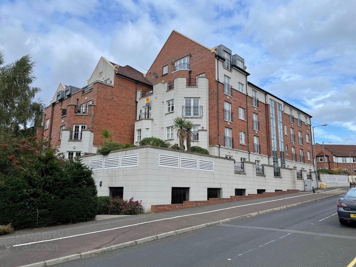 The building features a modern design with a mix of red brick and white stone. Lush greenery surrounds the entrance, while palm trees are positioned near the façade. A welcoming pedestrian pathway leads up to the front entrance, complemented by well-maintained landscaping.