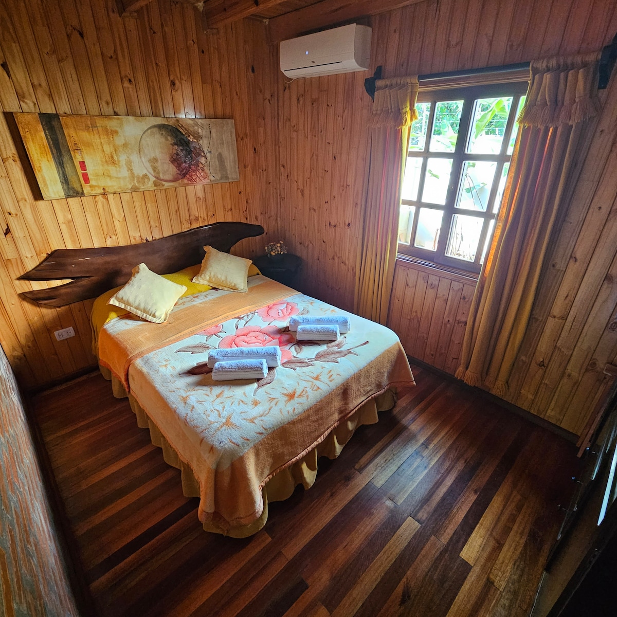 A rustic wooden room features a spacious bed adorned with a light yellow blanket and neatly arranged towels. Sunlight filters through the window, casting warmth on the wooden walls. Additional decorative elements include wall art and curtains complementing the natural tones.