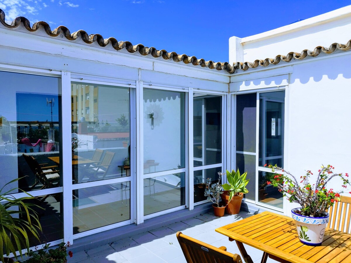A bright outdoor terrace bathed in sunlight is framed by large glass doors. A wooden table, accompanied by folding chairs, is positioned amongst potted plants. A tranquil atmosphere is complemented by the blue sky and the decorative tiles lining the terrace.