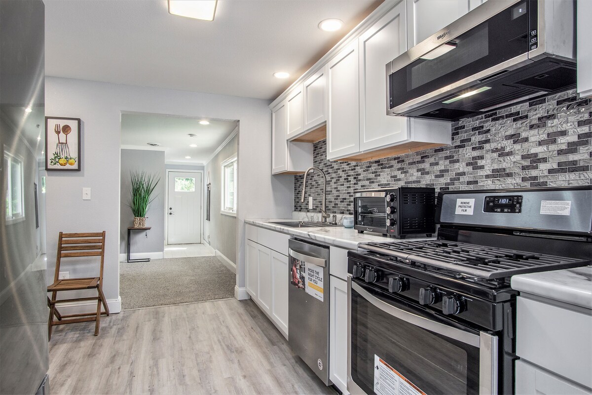 The fully stocked kitchen is equipped with a gas oven, stainless steel refrigerator, and microwave. Light wood cabinetry provides storage, while a gray tiled backsplash adds a contemporary touch. A wooden chair is positioned near the entrance, which leads to a well-lit hallway.