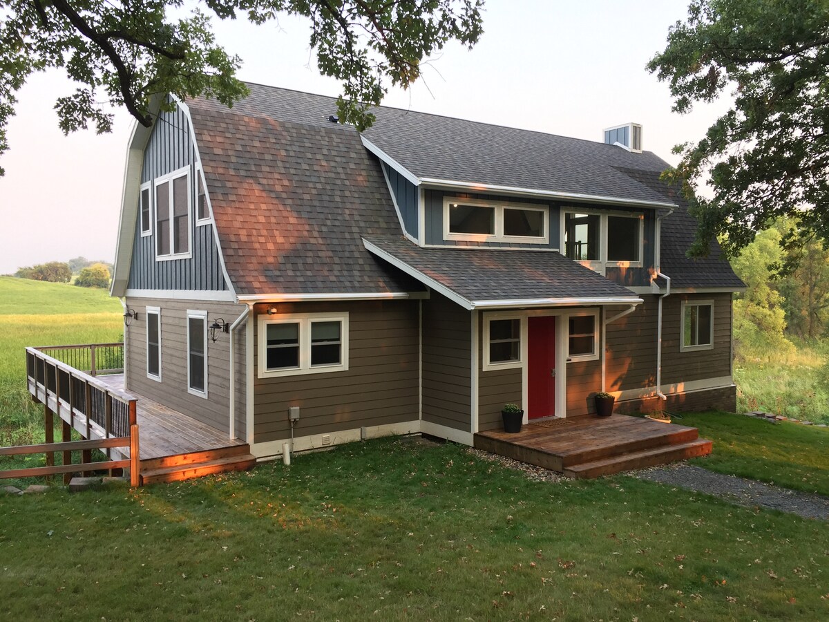A spacious country estate is depicted, featuring a combination of gray and natural wood siding. The front porch is framed by potted plants, and large windows allow for ample natural light. A deck extends from the front, overlooking a grassy area with distant hills.