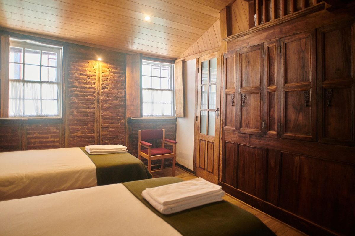 A cozy room features two single beds with green throws and neatly arranged towels. A wooden wardrobe stands against a rustic brick wall, while a chair is positioned beside a window, allowing natural light to brighten the space. A door to a private area is also visible.