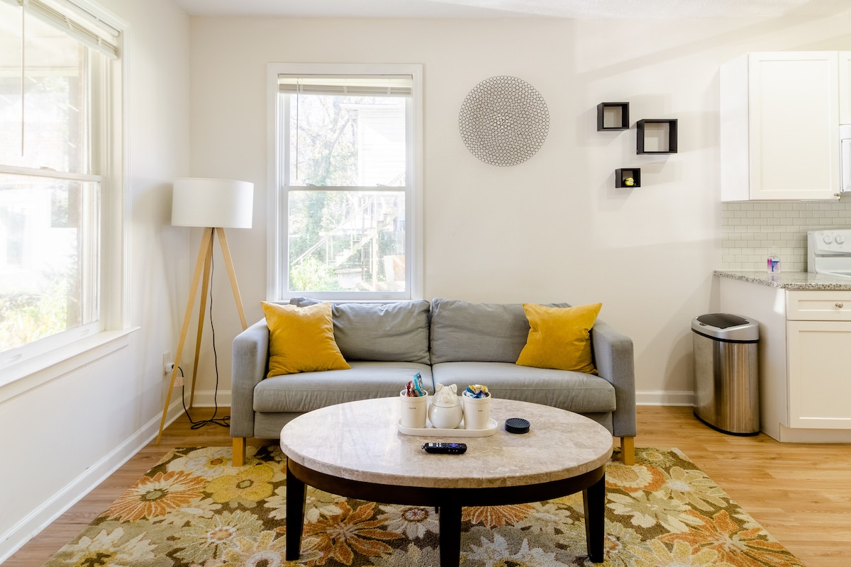 A cozy living area is presented with a light gray couch adorned with yellow accent pillows, positioned in front of a circular coffee table. Natural light enters from the two windows, enhancing the warm wood flooring. A kitchen area is visible in the background, equipped with white cabinetry.