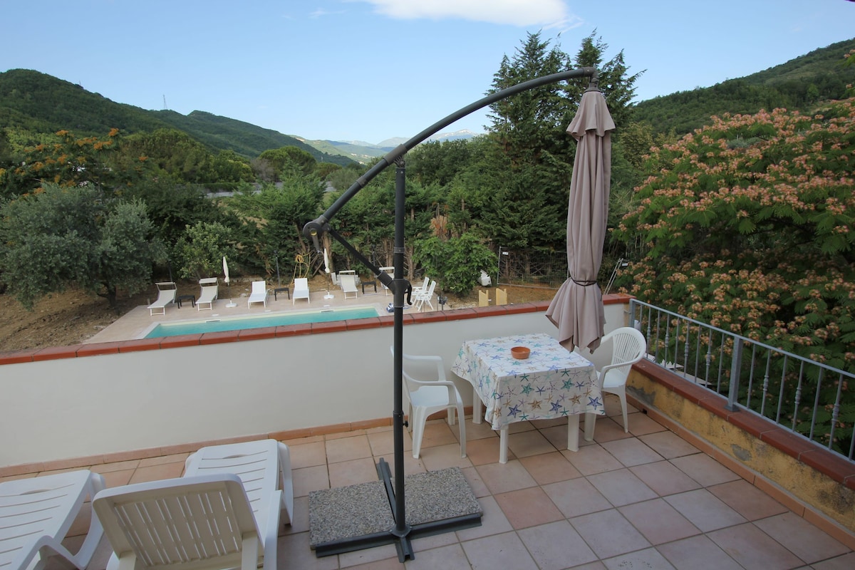 A terrace area is presented, featuring a table covered with a floral tablecloth. Two white chairs are positioned nearby, with a large umbrella on a stand providing shade. The view overlooks a swimming pool surrounded by lounge chairs, framed by lush greenery and mountainous terrain.