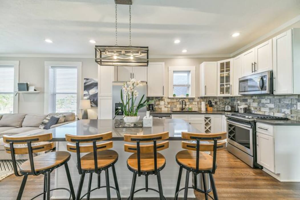 A modern kitchen is featured, showcasing stainless steel appliances and a stone backsplash. Four wooden barstools line the spacious countertop, complemented by a stylish light fixture overhead. Ample natural light pours in through nearby windows, enhancing the inviting atmosphere of the open space.