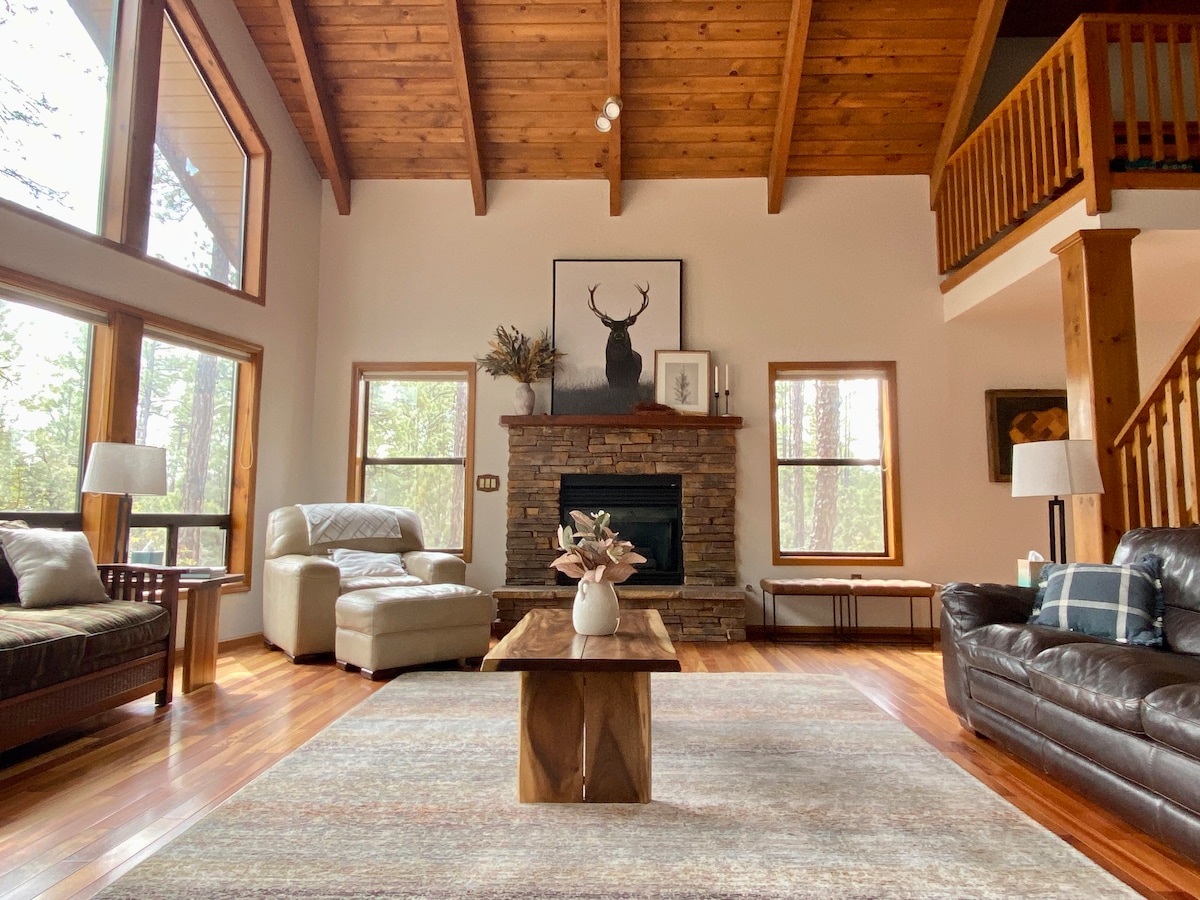 The living area showcases high cathedral ceilings with wooden beams, large windows allowing natural light, and a stone fireplace as a focal point. A coffee table sits at the center, surrounded by a plush seating arrangement, creating a welcoming space for relaxation.