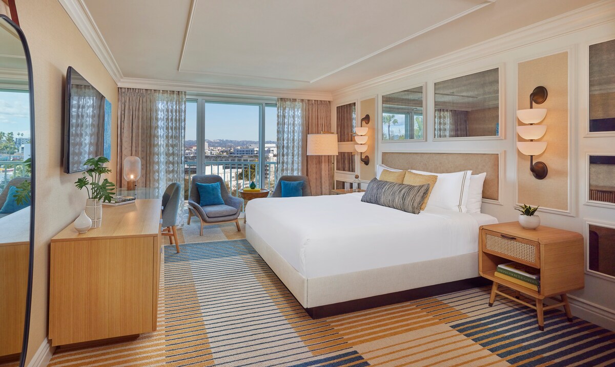 A spacious room features a large bed with a light-colored upholstered headboard and luxurious linens. Two armchairs in a vibrant hue sit nearby, alongside a stylish wooden dresser and a flat-screen TV. Floor-to-ceiling windows provide expansive city views and ample natural light.