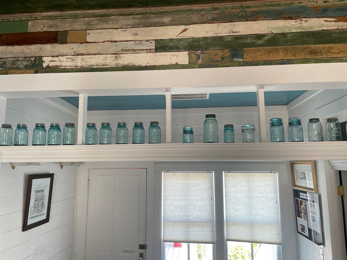 A row of vintage glass jars is displayed on a shelf above a doorway. The jars vary in size and shape, featuring a soft blue tint. Natural light filters in through two windows adorned with sheer window treatments, highlighting the rustic wooden wall behind.