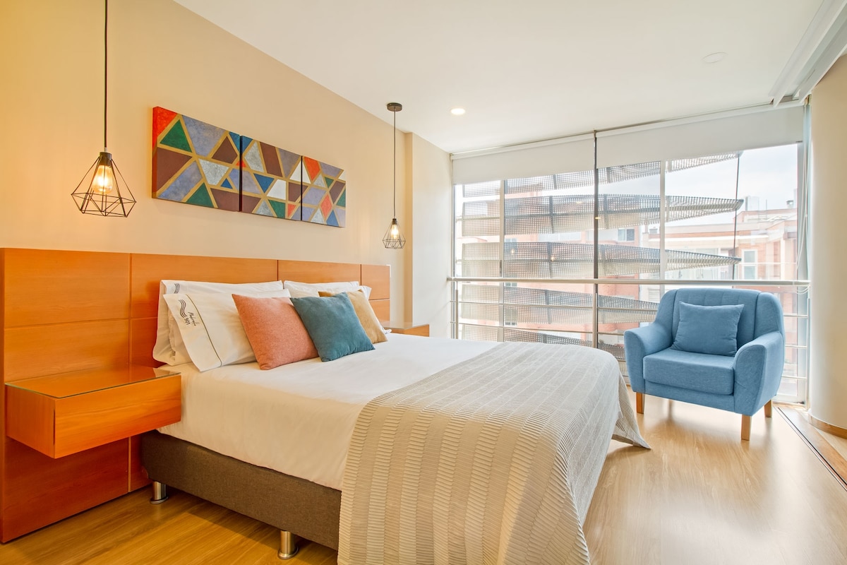 A modern bedroom features a spacious bed adorned with colorful pillows and a textured blanket. Ambient pendant lights hang above bedside tables. A cozy armchair in a soft blue color adds a pop of contrast. Large windows allow natural light to fill the room.