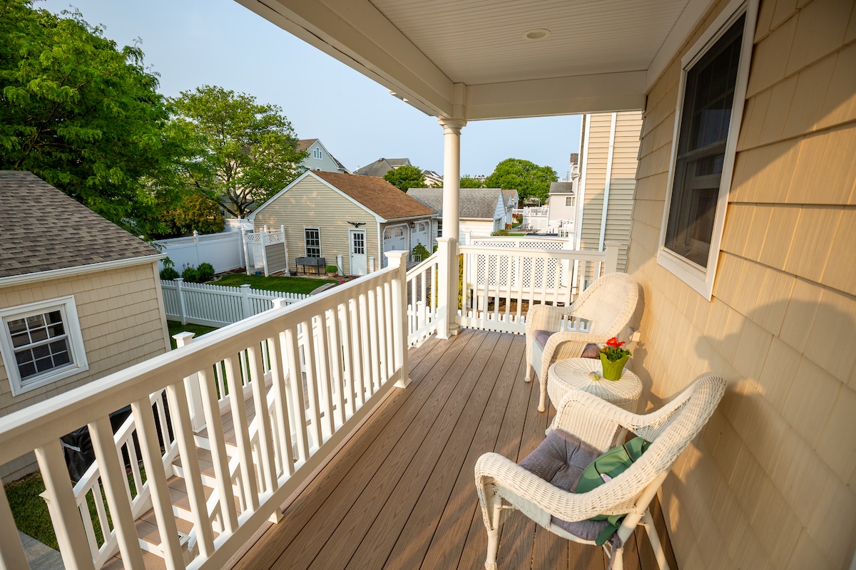 A covered balcony features two white wicker chairs positioned around a small round table. A potted plant adds a touch of greenery. The space is enclosed by white railings, offering a view of the neighboring yards with lush trees in the background.