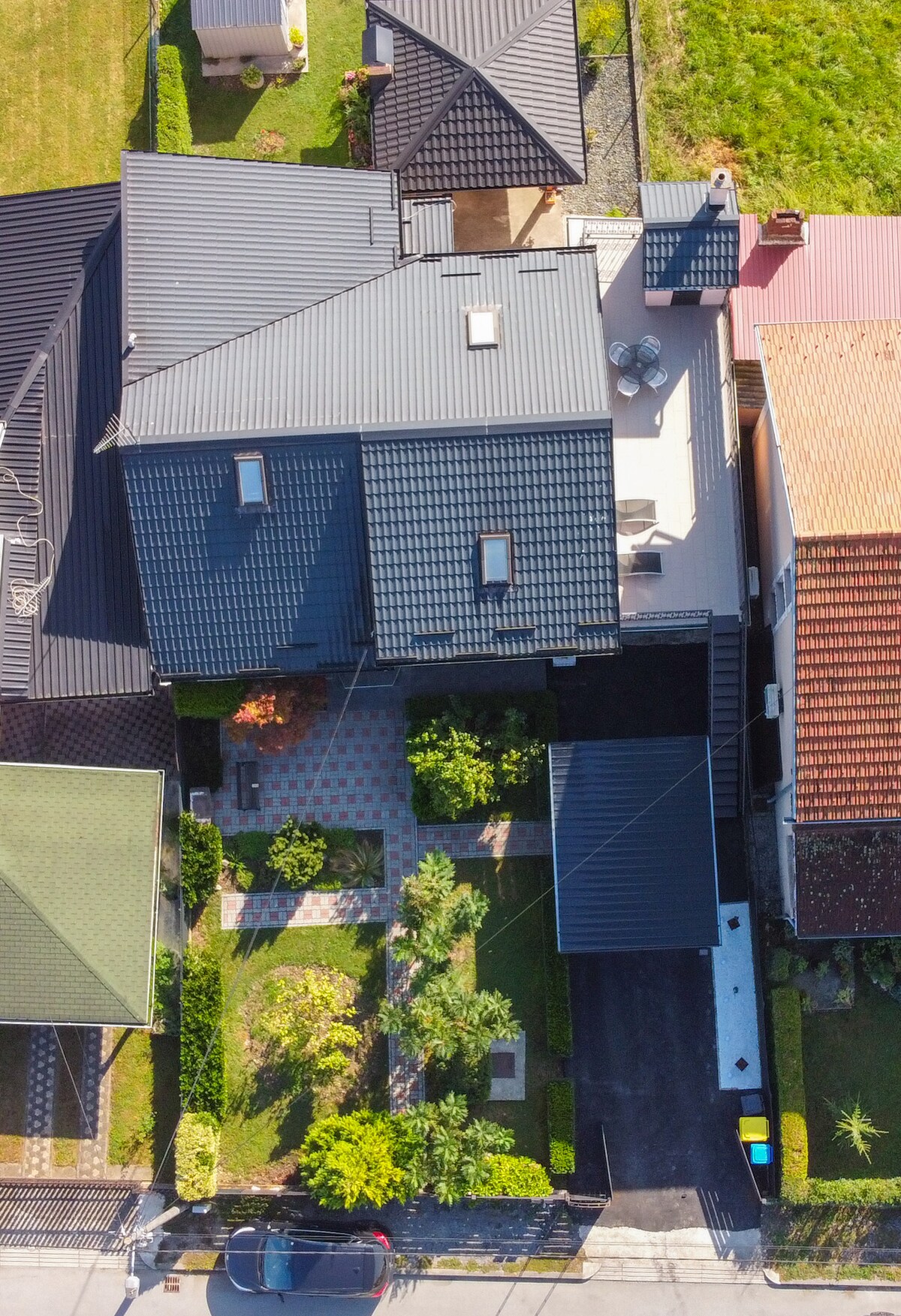 An aerial view of the property reveals a multi-level structure with a dark roof and landscaped gardens below. A spacious terrace is visible on the upper floor, while a well-maintained pathway leads through the greenery, which includes various shrubs and trees.