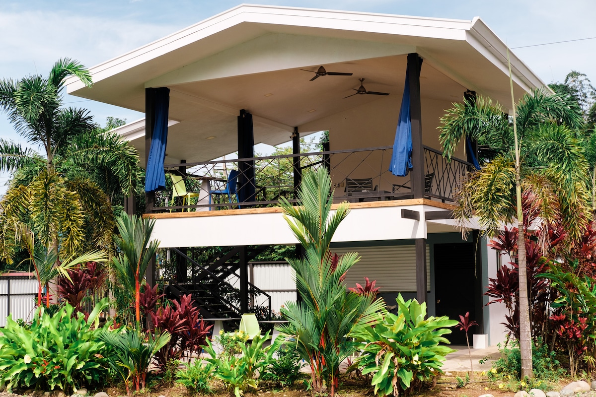 A two-story open air rancho is surrounded by lush tropical landscaping. The outdoor space features a shaded seating area with ceiling fans and vibrant blue curtains. Flourishing green plants and colorful foliage frame the structure, enhancing the inviting atmosphere.