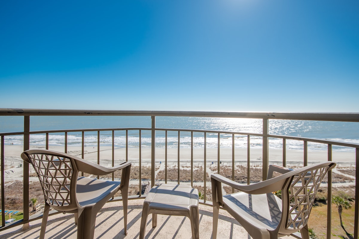 A balcony view showcases two light-colored chairs facing the ocean, with the beach and gently rolling waves visible below. The bright blue sky complements the serene scene, while sunlight reflects on the water's surface, enhancing the coastal atmosphere.