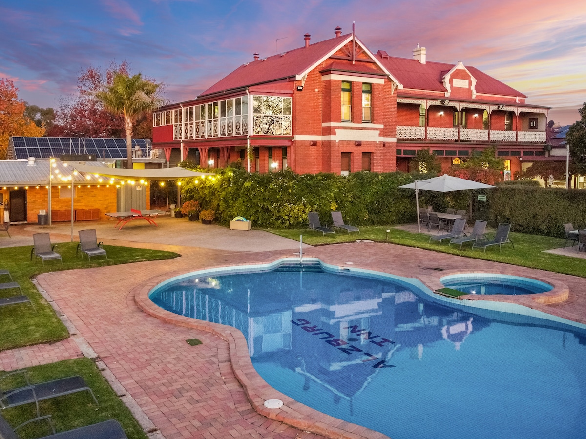A spacious outdoor pool area is surrounded by lounge chairs and festive lighting. The historic building with a red facade stands in the background, complemented by green shrubs and trees. The pool features the resort's name, reflecting the vibrant sunset colors in the sky.