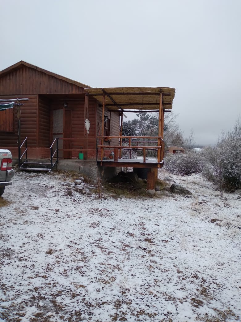 A rustic cabin is set in a snowy landscape, featuring a wooden porch with a thatched roof. The surrounding area is blanketed in white, with low shrubs and sparse vegetation visible. The entrance is accessed by a set of steps leading from the porch.