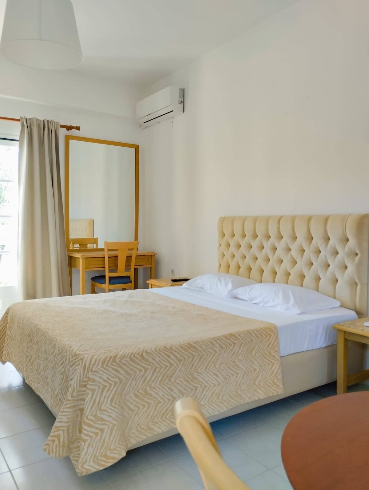 A serene bedroom is featured, showcasing a spacious bed with a soft, neutral-colored blanket. A mirrored dresser is positioned across from the bed, accompanied by a wooden chair. Natural light filters in through a nearby window, draped with light curtains.