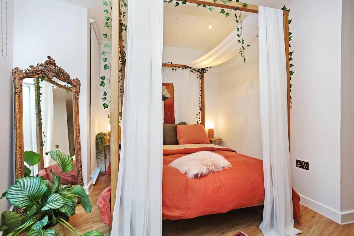 A four-poster king-sized bed is adorned with light drapery and earthy-toned bedding. A decorative mirror reflects the cozy design elements, while a lush green plant adds a touch of nature to the space. Soft lighting from bedside lamps creates a serene ambiance.