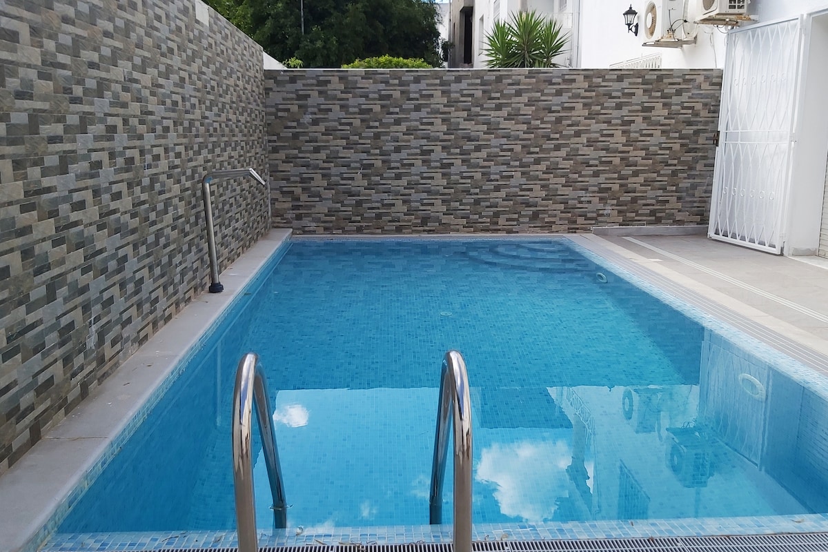 A private pool is showcased, featuring clear blue water and stainless steel stairs. The surrounding wall is decorated with stone tiles, providing a modern touch. Sunlight reflects gently on the water's surface, creating a serene atmosphere.
