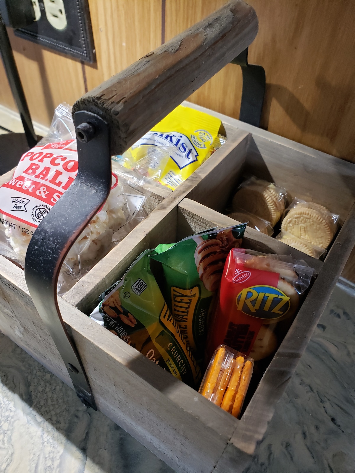 A rustic wooden container holds a variety of snacks, including popcorn, crackers, cookie packs, and cheese snacks. The container is divided into sections to neatly organize the items, and the surrounding area features wood paneling and a textured countertop.