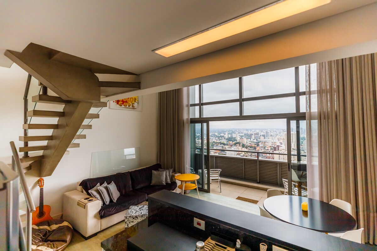 A modern living area is displayed, featuring a comfortable sectional sofa with accent pillows and a wooden coffee table. Large windows offer expansive city views, while a staircase leads to an upper level. The dining area is equipped with a round table and chairs.