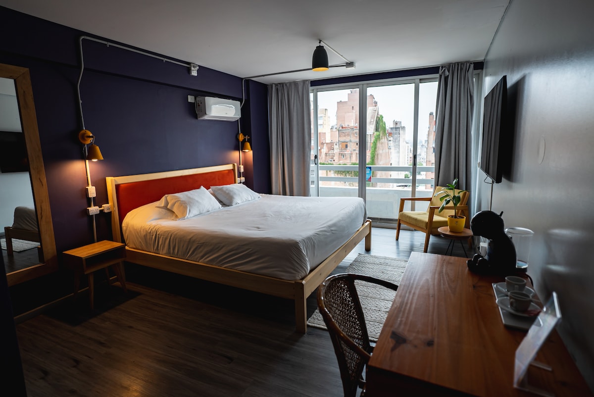A spacious room features a large bed against a deep blue wall. Natural light filters through floor-to-ceiling windows, revealing a view of the city. A desk with a chair and a coffee station are positioned near the window, complemented by a potted plant.