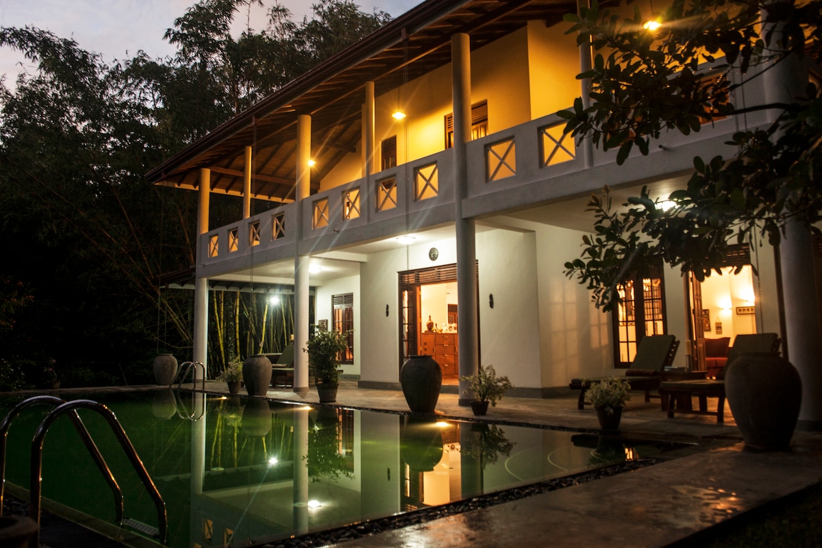 A two-story villa is illuminated by warm lighting, reflecting off the calm pool. The house's open design features large doors leading to the interior. Surrounding greenery adds to the serene atmosphere, enhancing the sense of privacy and tranquility.