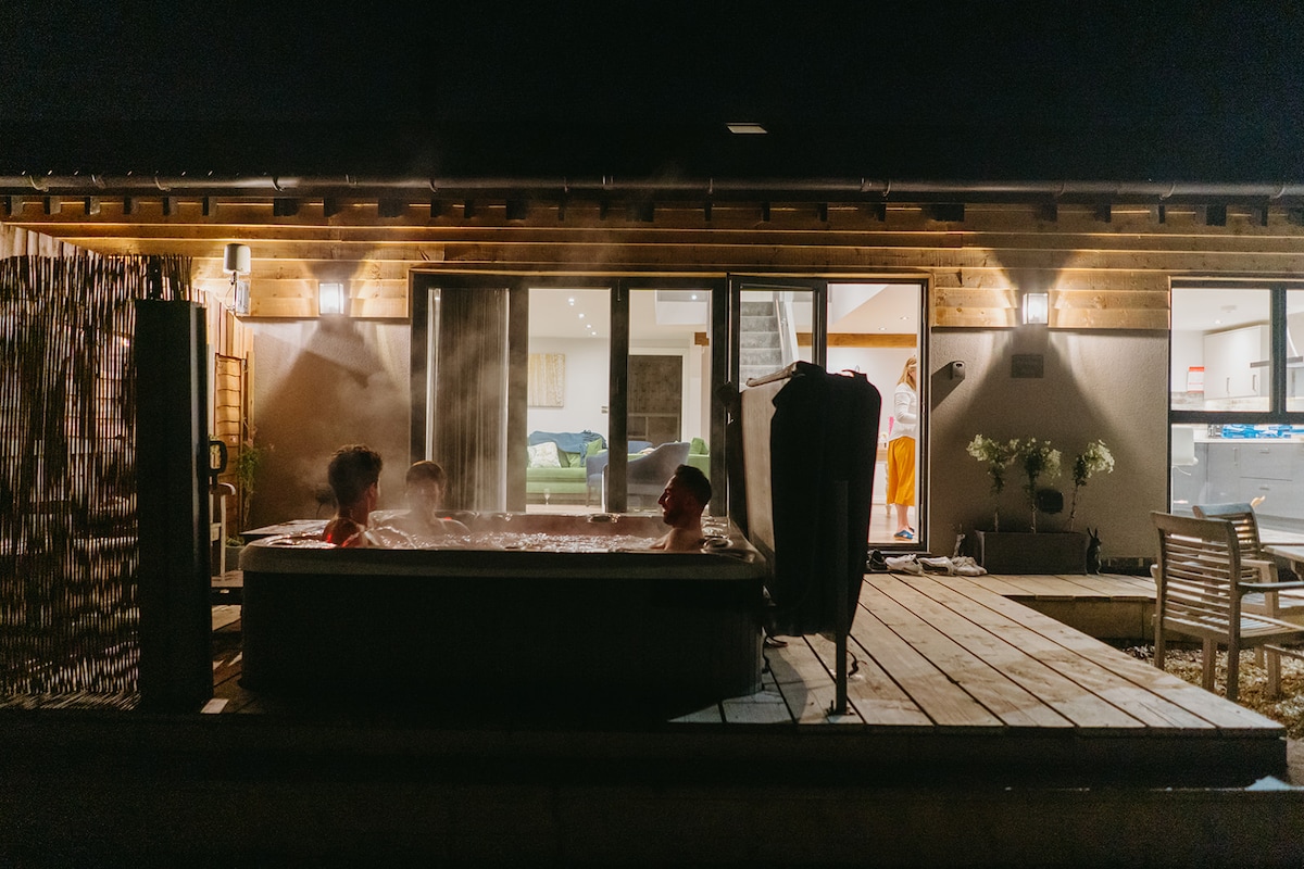 A hot tub for four sits on a wooden deck, emitting steam into the night air. Bi-fold doors lead into a well-lit interior. Soft lighting enhances the welcoming ambiance, creating a serene space for relaxation.