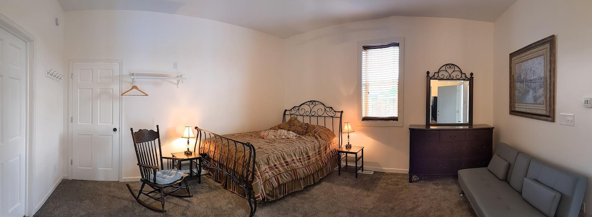 A spacious room is seen featuring a large metal-framed bed with patterned bedding. Two bedside lamps provide soft illumination, and a wooden rocking chair is positioned nearby. A mirror and a dresser occupy one wall, while a gray sofa extends along the opposite side.