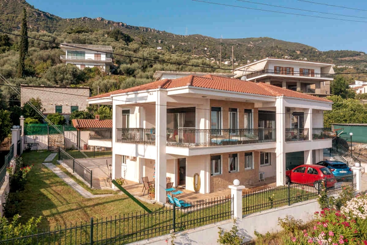 A modern two-storey house is showcased, featuring a red-tiled roof and spacious balconies. The surrounding lawn is well-maintained, complementing the nearby parking area with two cars. Green hills rise behind the property, creating a natural backdrop.