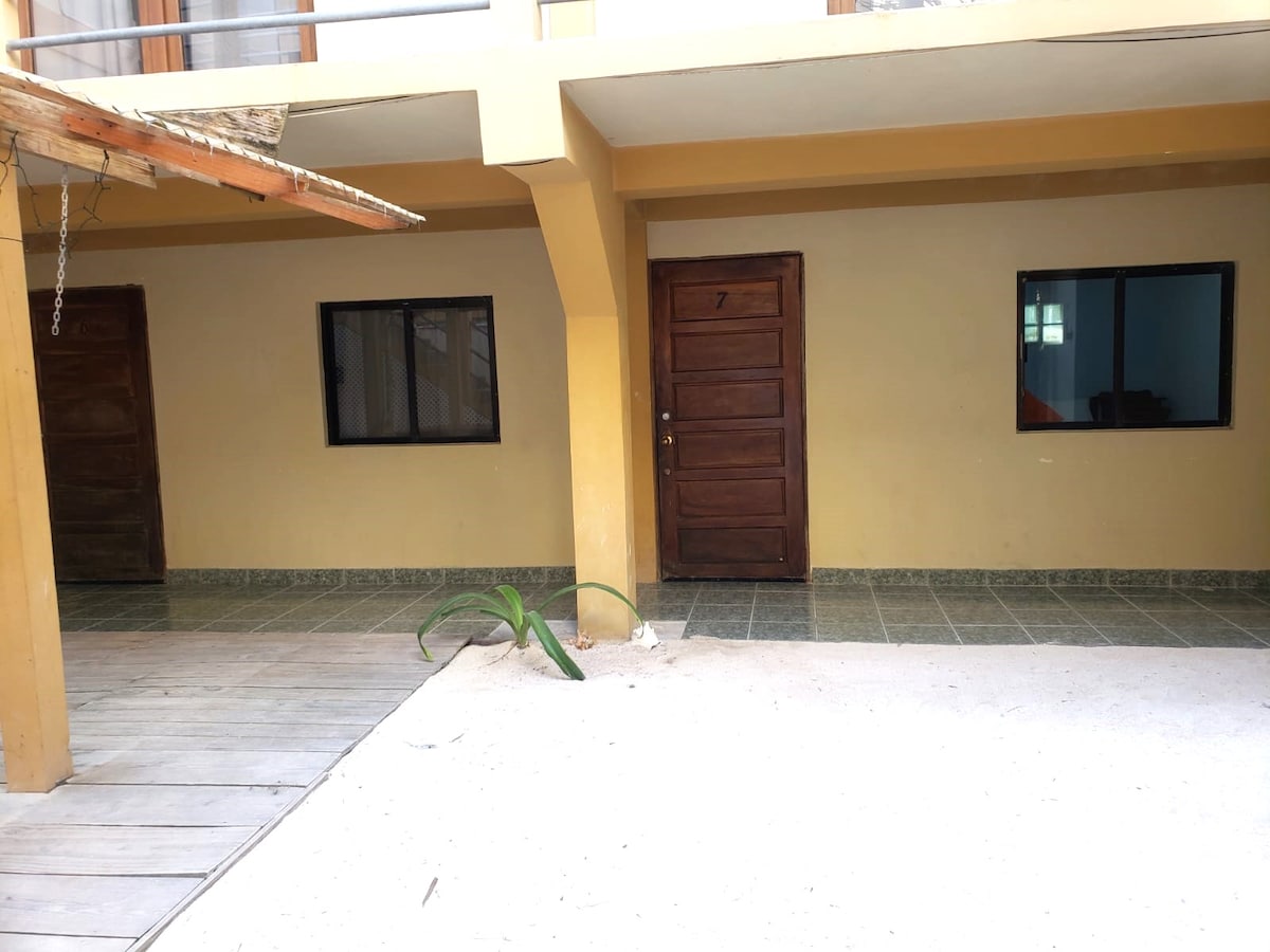 An outdoor view of the building's entrance showcases two private rooms with wooden doors. One window is visible for each room, allowing natural light to enter. A small green plant is seen on the sandy floor, contributing to the minimalistic layout of the space.