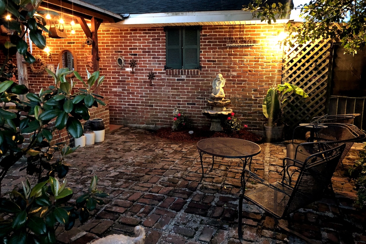 A brick patio is surrounded by greenery, featuring a small fountain as a centerpiece. A round table with chairs is positioned nearby, illuminated softly by overhead lights. The textured pavers create an inviting outdoor space, enhancing the ambiance for relaxation.