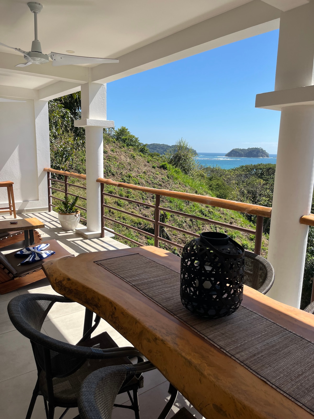 A spacious balcony features a large rustic dining table surrounded by comfortable chairs, offering views of the ocean and lush greenery. A decorative lantern rests centrally on the table, while the coastline is visible in the distance under a clear blue sky.