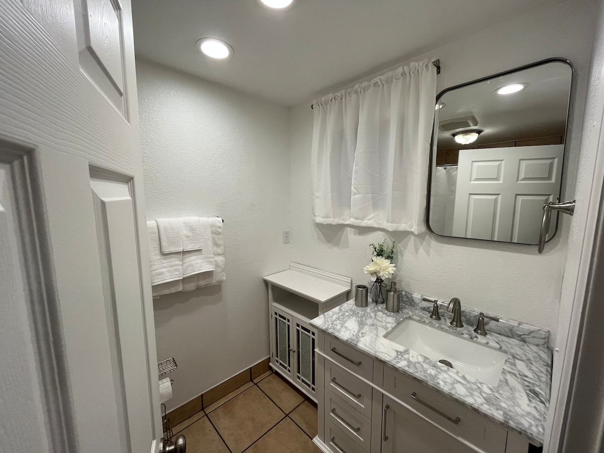 A clean and modern bathroom is showcased, featuring a marble countertop and a large mirror. Soft, white towels are neatly hung on the wall, while a small vase of fresh flowers adds a touch of nature. The space is well-lit, enhancing the bright atmosphere.