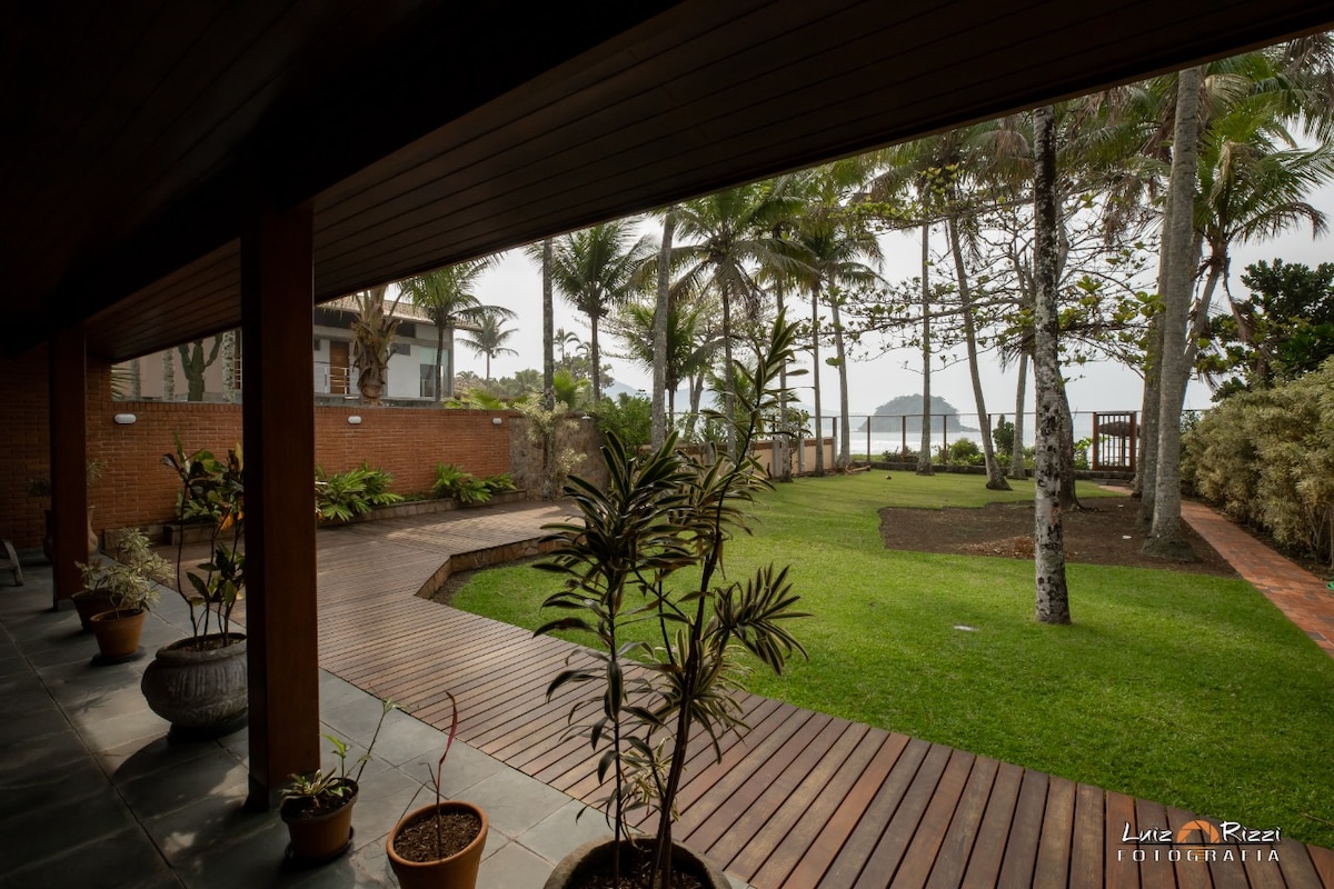 A view from a covered terrace shows a landscaped garden with lush grass and palm trees. An inviting wooden deck leads to the beach, creating a seamless connection to the outdoor space. The distant shoreline can be seen through the greenery.