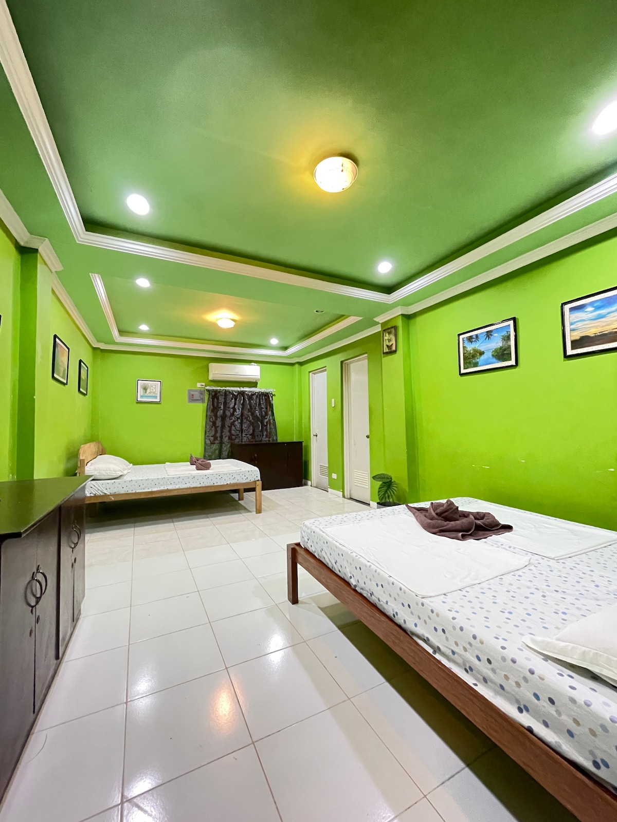 A spacious guest room is decorated with vibrant green walls and features two unmade beds with simple bedding. A wooden dresser is positioned against one wall, while framed pictures adorn the other walls. Soft lighting creates an inviting atmosphere, complemented by a small window with curtains.