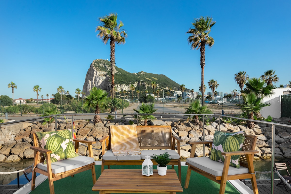A modern outdoor terrace features a seating area with two wooden chairs adorned with green leaf-patterned cushions and a coffee table. Lush palm trees surround the space, with a view of a rocky mountain in the background under clear blue skies.