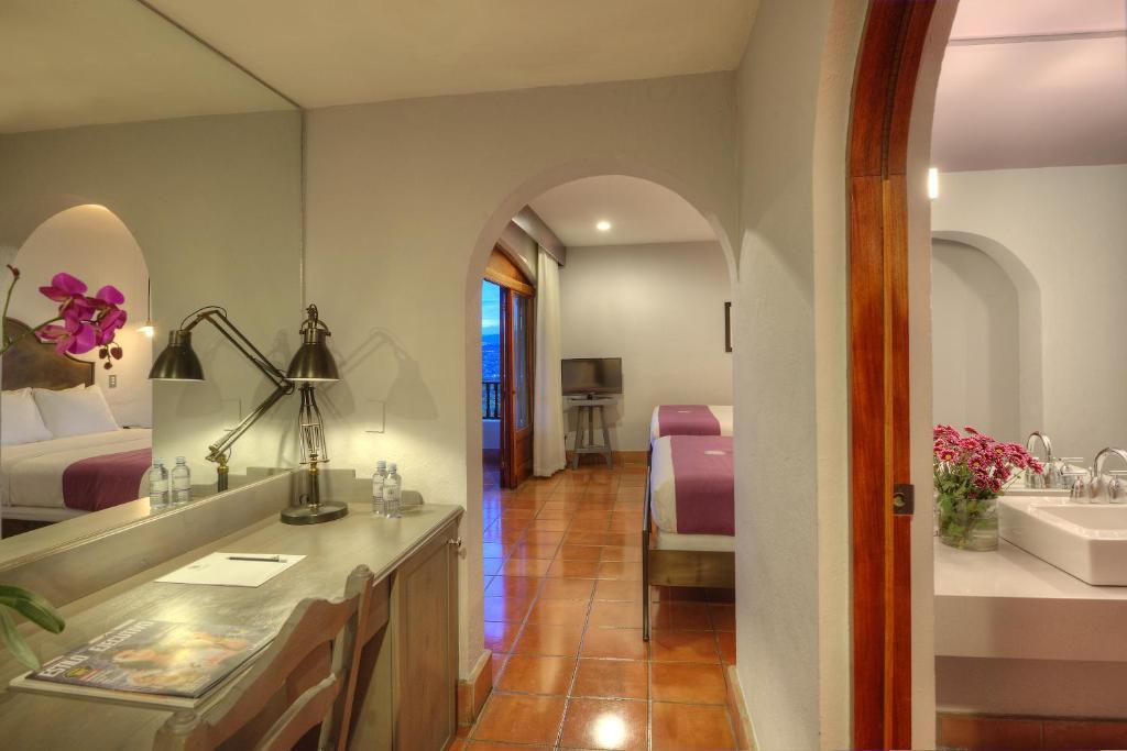 An inviting room layout is displayed, featuring two queen beds with elegant bedding. A desk with a lamp sits beside a window, while a television is visible across the room. Soft lighting highlights the earthy tile floor and colorful floral arrangement on the countertop.