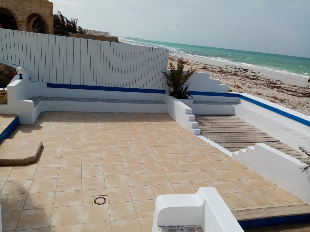 A spacious terrace features light-colored tiles, bordered by smooth white walls and splashes of blue. Steps lead down to the beach, while palm plants are placed decoratively. The expansive ocean view is visible beyond, highlighting the serene beachfront setting.