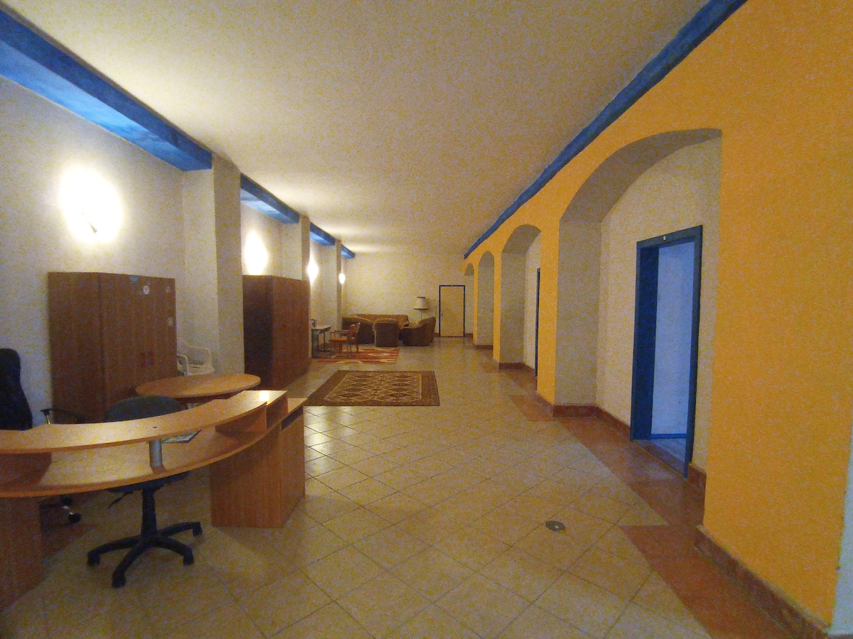 A spacious interior features a combination of light-colored walls and blue accents. A large reception desk is positioned to the left, with comfortable seating arranged in the background. Arched doorways and warm lighting create an open and inviting environment.