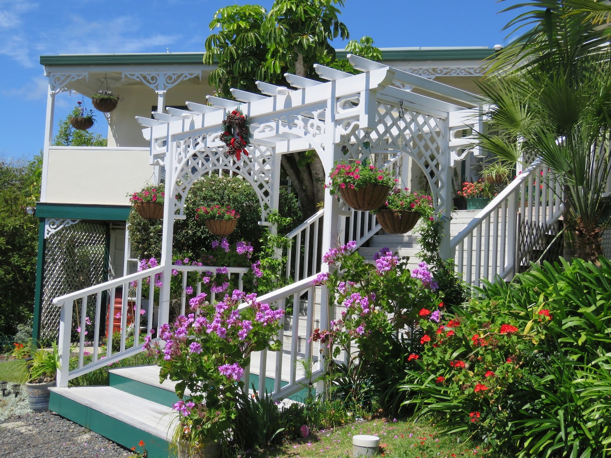 A charming entrance is framed by a white trellis adorned with hanging planters, featuring vibrant blooms in shades of pink and purple. Lush greenery surrounds the steps leading up to the cottage, creating a welcoming atmosphere in a bright, sunny setting.