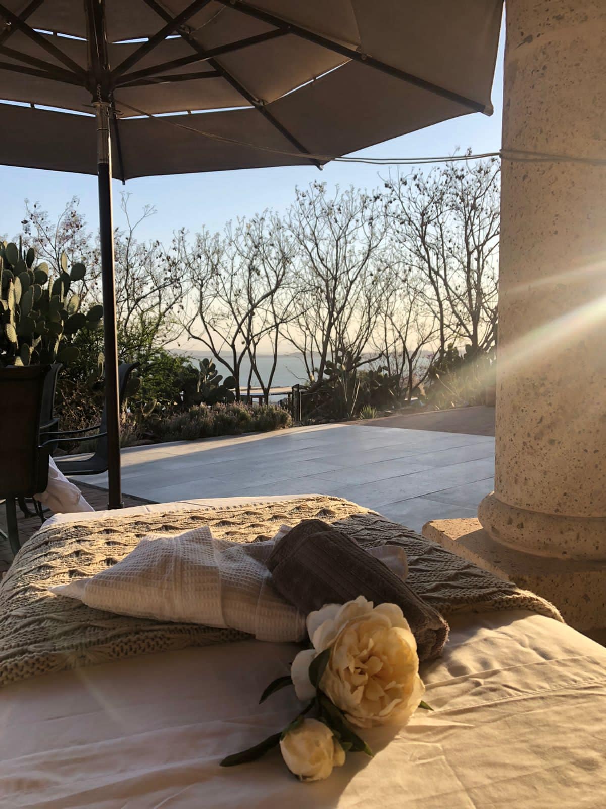 A sunlit outdoor seating area features a cozy lounge chair adorned with a soft blanket. A white flower rests gently on the fabric, while a sun umbrella provides shade. The background reveals trees and distant hills, enhancing the serene atmosphere.