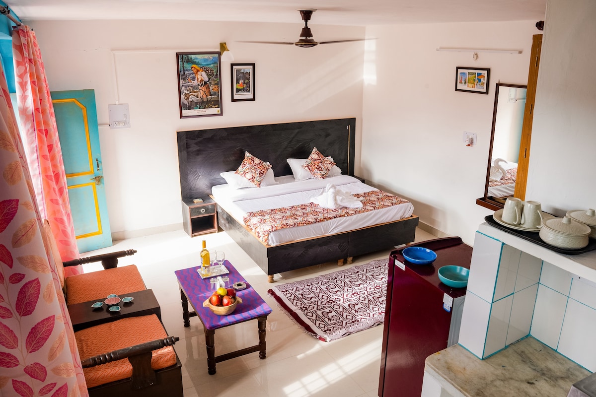 A spacious room is presented with a double bed adorned with decorative pillows. A mirror reflects the room's furnishings, including a red refrigerator and a kitchenette area. Natural light filters through curtains, while a small table holds fruits and drinks, adding convenience for guests.