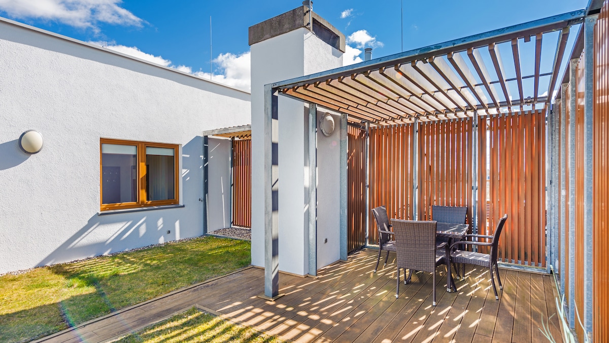 An outdoor terrace is featured, surrounded by wooden slats for privacy. A table with four chairs is positioned on a wooden deck, complemented by a small patch of grass. The terrace is bathed in natural light, creating a serene ambiance.