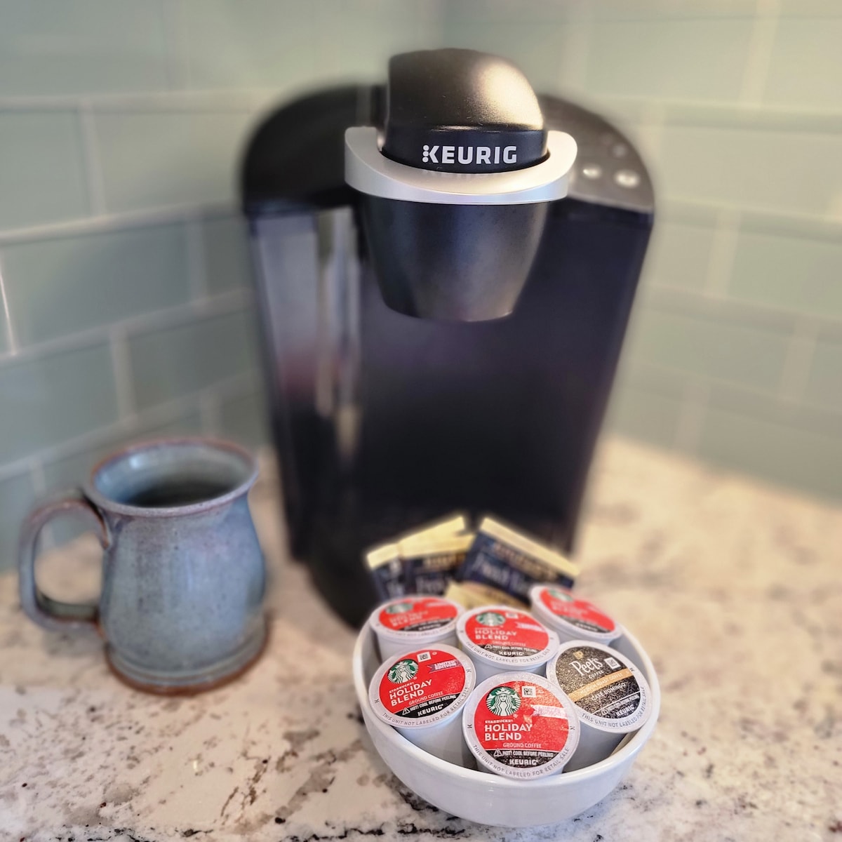 A Keurig coffee maker is positioned against a light green tile backsplash. A bowl containing assorted coffee pods is placed on a countertop, alongside a ceramic coffee mug filled with dark liquid. Additional coffee pods are visible in the background.
