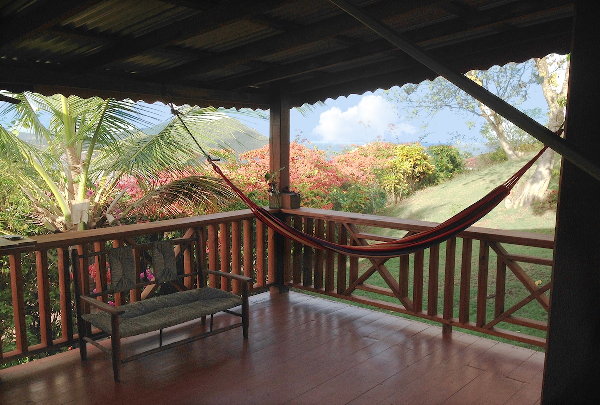 A covered veranda features a colorful hammock suspended between wooden posts alongside a woven seating arrangement. Lush greenery and flowering plants are visible in the background, creating a peaceful outdoor setting under a partially cloudy sky.