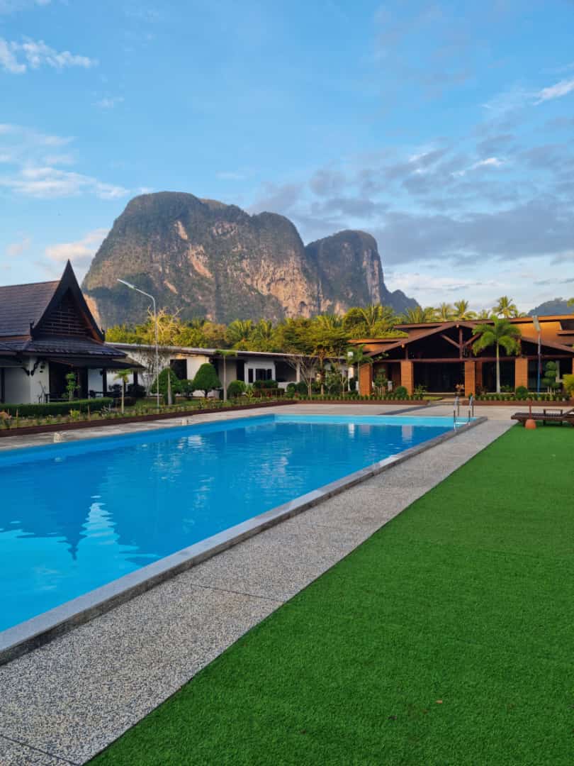 A rectangular swimming pool is bordered by a manicured lawn and stone pathway, with a backdrop of dramatic mountains. Nearby structures feature traditional architectural designs, and palm trees provide a touch of greenery, contributing to a relaxing environment.